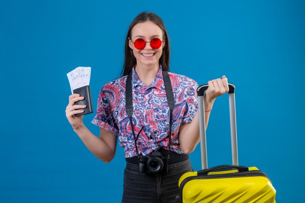 Giovane viaggiatore donna che indossa occhiali da sole rossi in piedi con la valigia gialla tenendo il passaporto e biglietti sorridendo allegramente guardando la fotocamera con la faccia felice su sfondo blu
