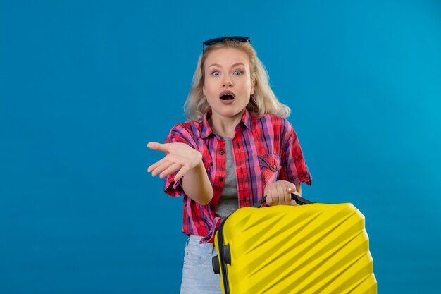 Giovane viaggiatore femminile spaventato che indossa camicia rossa e occhiali sulla testa che tiene i punti della valigia a lato sulla parete blu isolata