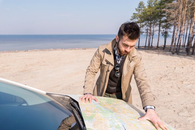 Giovane viaggiatore maschio che cerca la posizione sulla mappa sopra il cofano dell&#39;automobile alla spiaggia