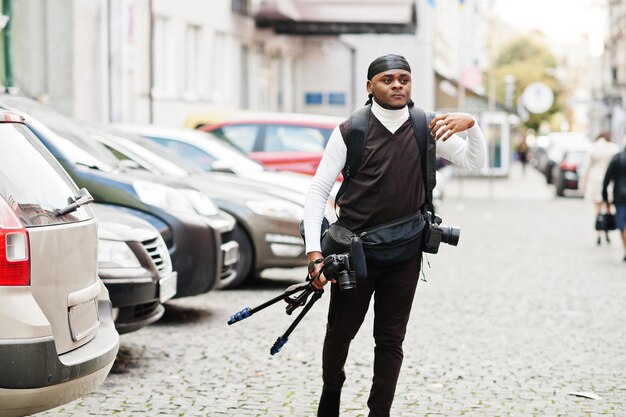 Giovane videografo afroamericano professionista che tiene una fotocamera professionale con attrezzatura pro treppiede Cameraman afro che indossa duraq nero che realizza un video