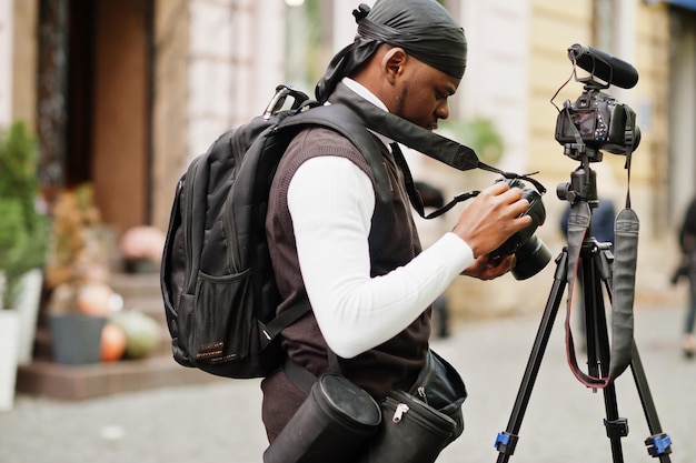 Giovane videografo afroamericano professionista che tiene una fotocamera professionale con attrezzatura pro treppiede Cameraman afro che indossa duraq nero che realizza un video