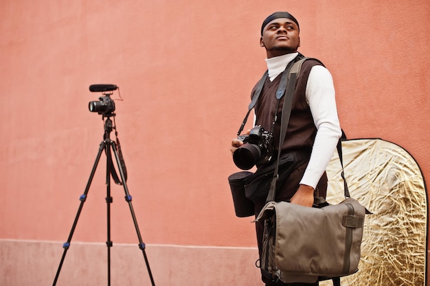 Giovane videografo afroamericano professionista che tiene una fotocamera professionale con attrezzatura professionale Cameraman afro che indossa un duraq nero che realizza un video