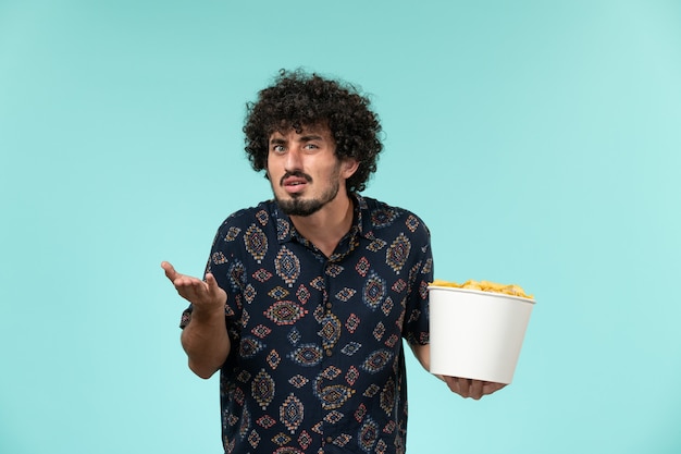 Giovane vista frontale che tiene il canestro con le patatine fritte su un teatro cinematografico della pellicola di film a distanza della parete blu