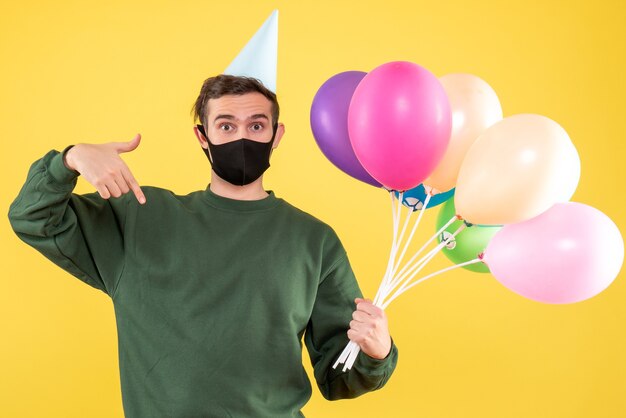 Giovane vista frontale con cappello da festa e palloncini colorati che punta con il dito verso il basso in piedi sul giallo