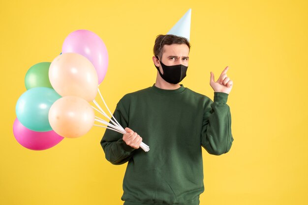 Giovane vista frontale con cappello da festa e palloncini colorati che punta sul retro in piedi sul giallo