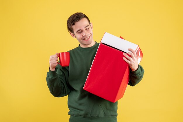 Giovane vista frontale con maglione verde che tiene grande regalo e tazza rossa in piedi sul giallo