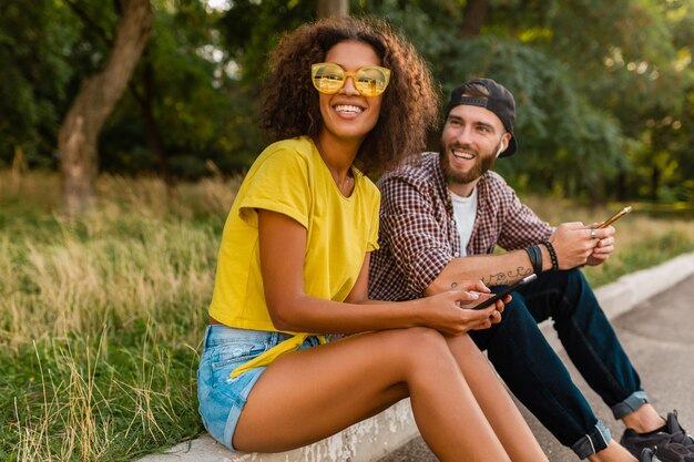 Giovani amici sorridenti felici che si siedono parco utilizzando smartphone, uomo e donna che hanno divertimento insieme