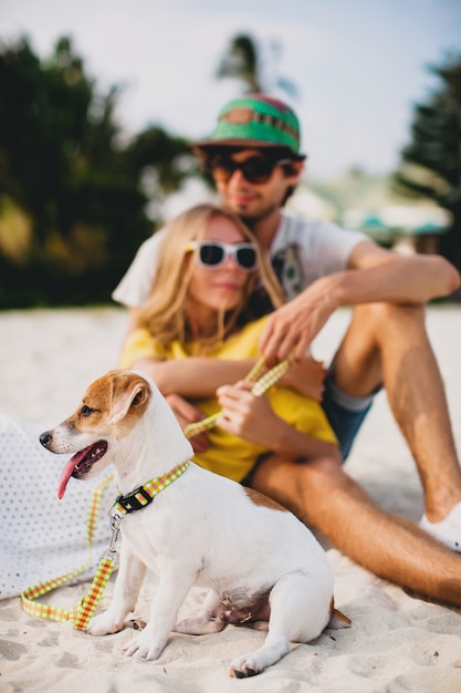 Giovani coppie alla moda hipster innamorate che camminano e giocano con il cane in spiaggia tropicale, abbigliamento cool, stato d'animo romantico, divertimento, sole, uomo donna insieme, orizzontale, vacanza, casa casa villa