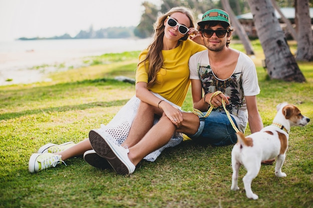 Giovani coppie alla moda hipster nell'amore che si siede sull'erba che gioca con il cane in spiaggia tropicale