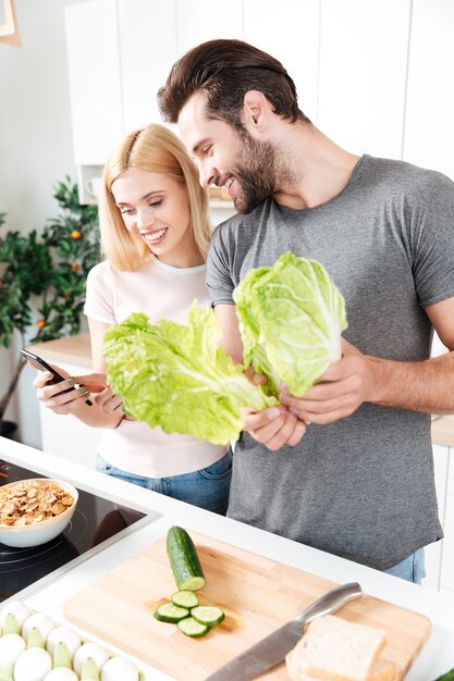 Giovani coppie amorose che cucinano insieme facendo uso del telefono cellulare