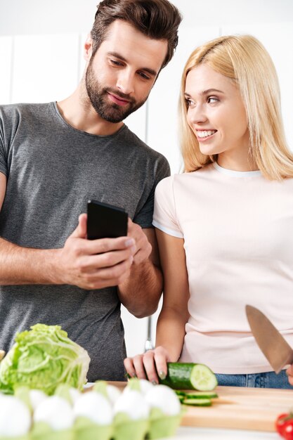 Giovani coppie amorose felici che cucinano insieme facendo uso del telefono