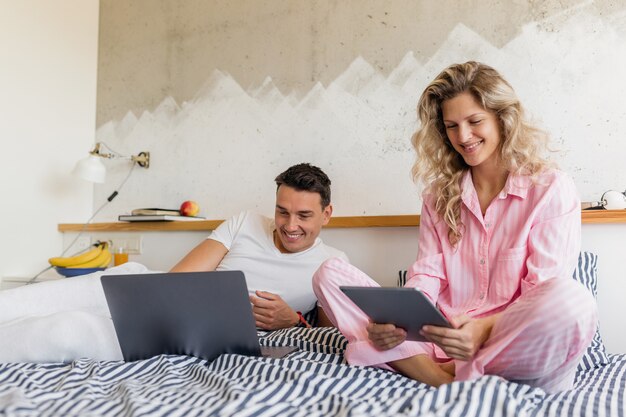 Giovani coppie attraenti che si siedono sul letto la mattina, leggendo le notizie in internet, lavoro libero professionista online