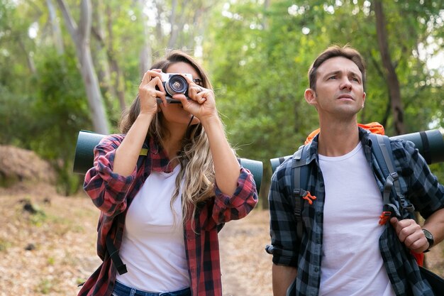 Giovani coppie caucasiche che fanno un'escursione nella foresta e che prendono la foto con la macchina fotografica. Riflessivo viaggiatore maschio in piedi vicino a donna e guardando il paesaggio. Concetto di turismo, avventura e vacanze estive con lo zaino in spalla