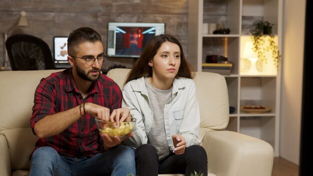 Giovani coppie caucasiche che mangiano patatine mentre si guarda la televisione. Coppia in cerca preoccupata per la televisione.