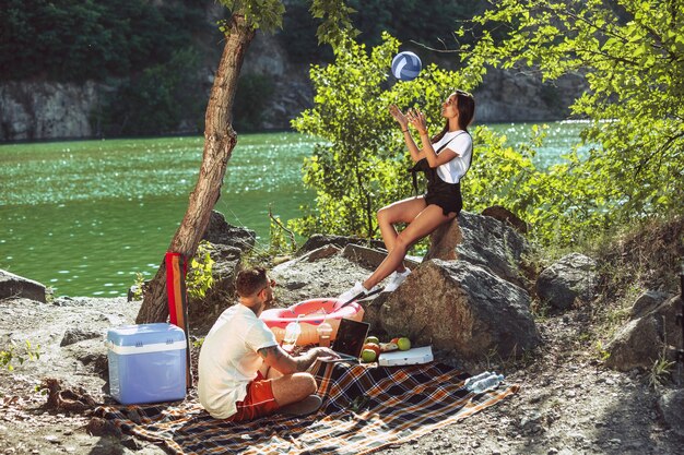 Giovani coppie che hanno picnic in riva al fiume in una giornata di sole. Donna e uomo che trascorrono del tempo insieme sulla natura. Divertirsi, mangiare, giocare e ridere. Concetto di relazione, amore, estate, fine settimana.