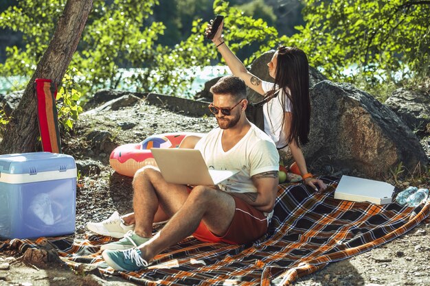 Giovani coppie che hanno picnic in riva al fiume in una giornata di sole. Donna e uomo che trascorrono del tempo sulla natura insieme. Divertirsi, mangiare, giocare e ridere. Concetto di relazione, amore, estate, fine settimana.