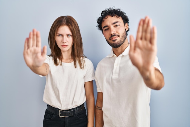 Giovani coppie che indossano abiti casual in piedi insieme facendo smettere di cantare con il palmo della mano. espressione di avvertimento con gesto negativo e serio sul viso.