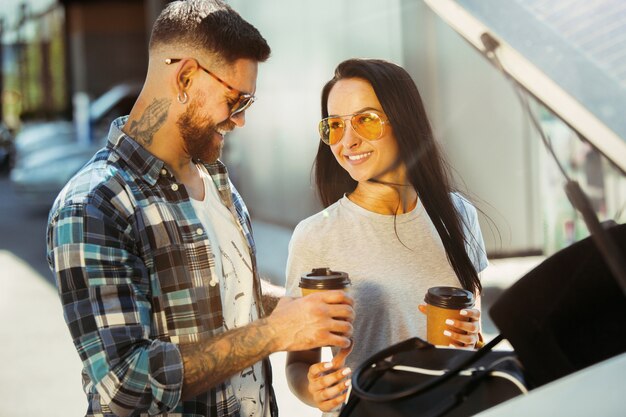 Giovani coppie che preparano per il viaggio di vacanza in macchina in una giornata di sole. Donna e uomo che bevono caffè e pronti per andare al mare o all'oceano.