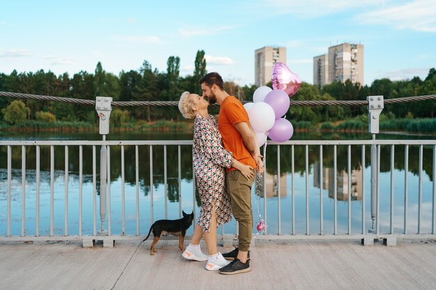 Giovani coppie felici che abbracciano e baciano sul ponte che tiene palloncini rosa e cane con la città all'orizzonte