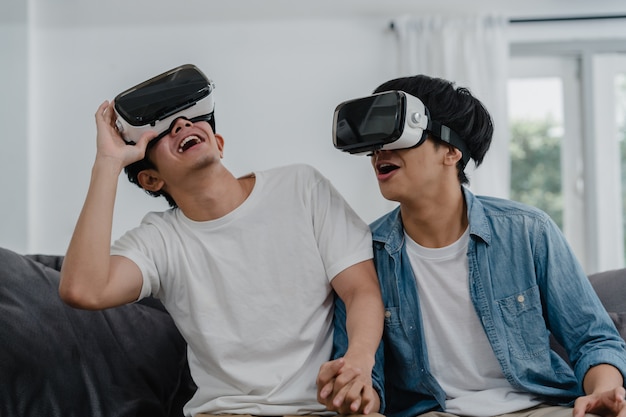 Giovani coppie gay asiatiche che utilizzano la tecnologia divertente a casa, ragazzo amante dell'Asia lgbtq + sentirsi felici divertimento e realtà virtuale, VR che giocano insieme mentre giace il divano nel salotto di casa.