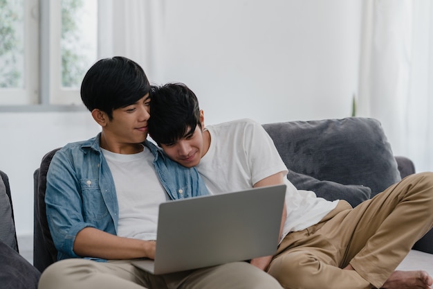 Giovani coppie gay facendo uso del computer portatile del computer a casa moderna. Gli uomini asiatici LGBTQ + si rilassano felici usando la tecnologia guardando film in internet insieme mentre giacciono sul divano nel salotto di casa.