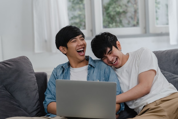 Giovani coppie gay facendo uso del computer portatile del computer a casa moderna. Gli uomini asiatici LGBTQ + si rilassano felici usando la tecnologia guardando film in internet insieme mentre giacciono sul divano nel salotto di casa.