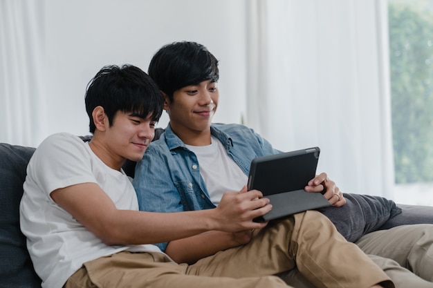 Giovani coppie gay facendo uso della compressa a casa. Gli uomini asiatici LGBTQ + si rilassano felici usando la tecnologia guardando film in internet insieme mentre giacciono sul divano nel soggiorno.