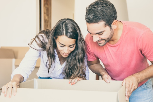 Giovani coppie latine eccitate felici che aprono la scatola di cartone e guardano dentro, spostano e disimballano le cose