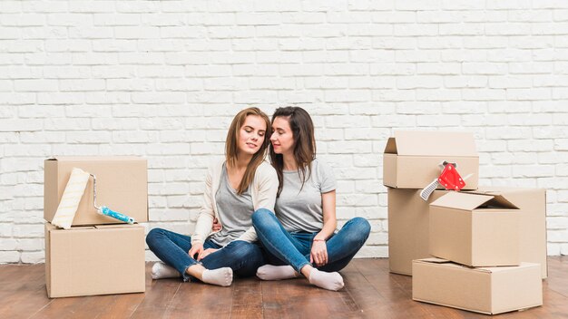 Giovani coppie lesbiche amorose che si siedono sul pavimento di legno duro con le scatole di cartone commoventi nella loro nuova casa