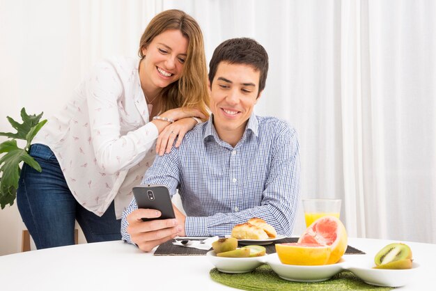 Giovani coppie sorridenti che esaminano telefono cellulare al tavolo della prima colazione