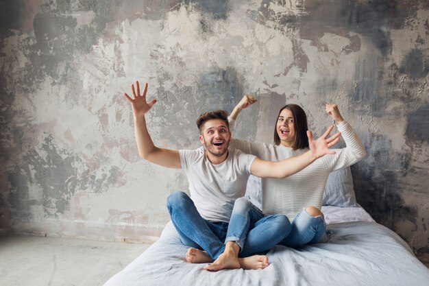 Giovani coppie sorridenti che si siedono sul letto a casa in abbigliamento casual, uomo e donna che hanno divertimento insieme, folle emozione positiva, felice, alzando le mani