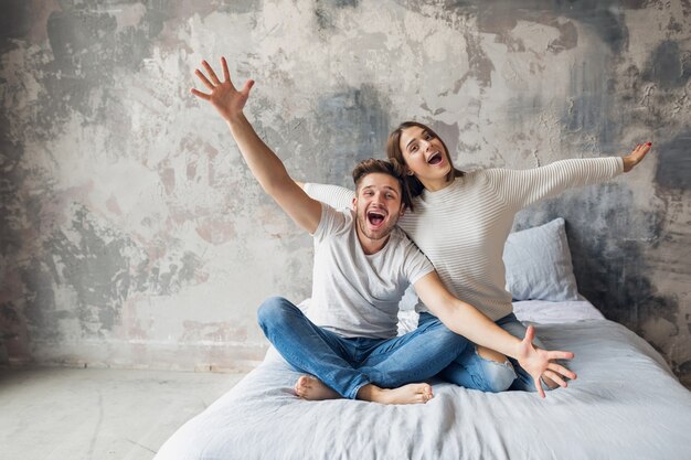 Giovani coppie sorridenti che si siedono sul letto a casa in abbigliamento casual, uomo e donna divertendosi insieme, folle emozione positiva, felice, alzando le mani