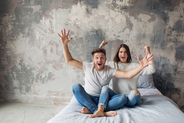 Giovani coppie sorridenti che si siedono sul letto a casa in abbigliamento casual, uomo e donna divertendosi insieme, folle emozione positiva, felice, alzando le mani