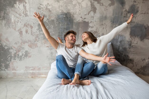 Giovani coppie sorridenti che si siedono sul letto a casa in abbigliamento casual, uomo e donna divertendosi insieme, folle emozione positiva, felice, alzando le mani