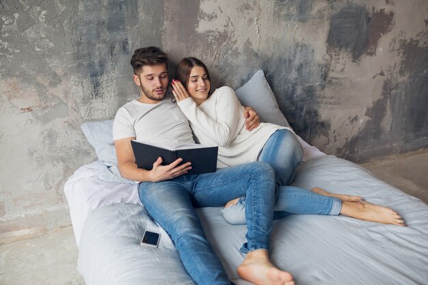 Giovani coppie sorridenti felici che si siedono sul letto a casa nel libro di lettura dell'abbigliamento casual che indossa jeans, uomo e donna che trascorrono del tempo romantico insieme