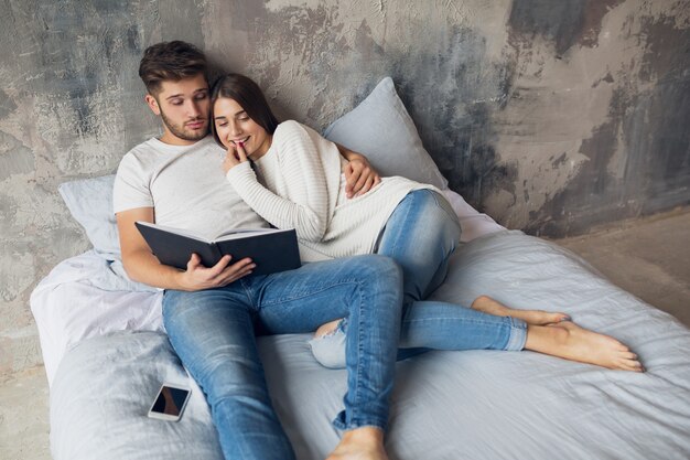 Giovani coppie sorridenti felici che si siedono sul letto a casa nel libro di lettura dell'abbigliamento casual che indossa jeans, uomo e donna che trascorrono del tempo romantico insieme