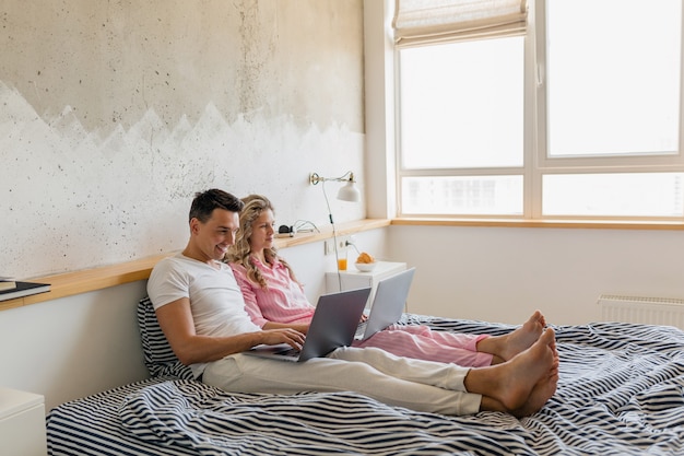 Giovani coppie sveglie che si siedono sul letto mattina, uomo e donna che lavorano al computer portatile