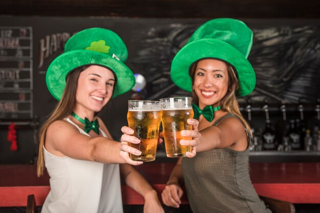 Giovani donne che ridono in cappelli di Saint Patricks mostrando bicchieri di drink al bancone del bar