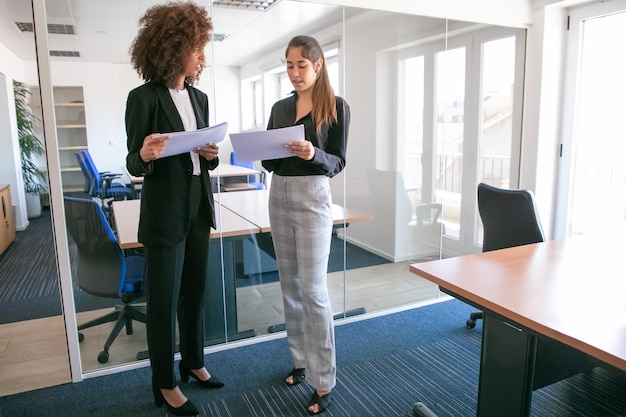 Giovani donne di affari attraenti che discutono della documentazione nelle mani. Due colleghe piuttosto fiduciose in possesso di documenti e in piedi nella stanza dell'ufficio. Concetto di lavoro di squadra, affari e gestione