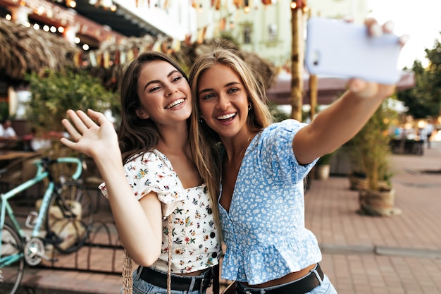 Giovani donne felici in eleganti camicette floreali sorridono sinceramente e si fanno selfie all'aperto