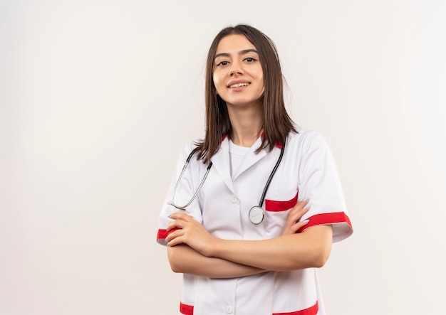 Giovani donne medico in camice bianco con lo stetoscopio intorno al collo guardando in avanti con la mano sul petto guardando fiducioso in piedi sul muro bianco
