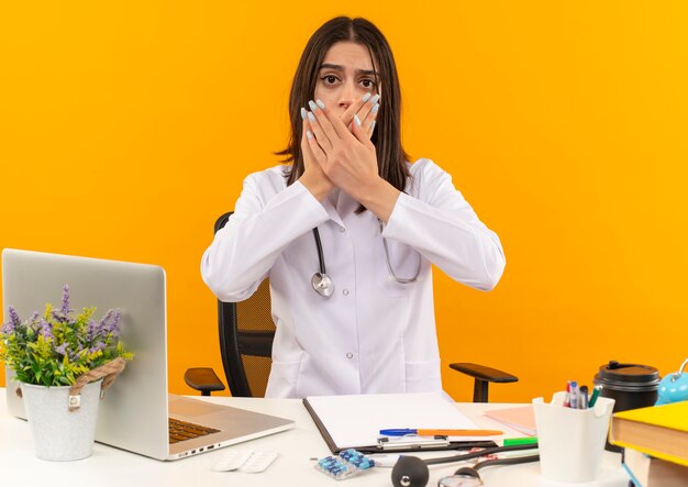 Giovani donne medico in camice bianco con uno stetoscopio guardando in avanti scioccato che copre la bocca con le mani seduto al tavolo con laptop e documenti sulla parete arancione