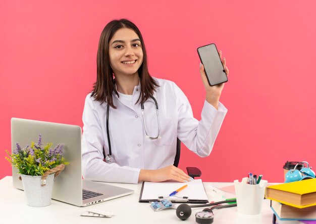 Giovani donne medico in camice bianco con uno stetoscopio intorno al collo che mostra lo smartphone sorridente fiducioso seduto al tavolo con il computer portatile sulla parete rosa