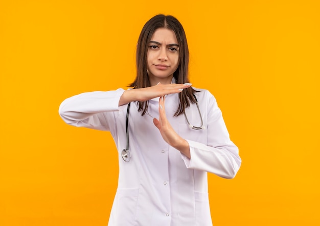 Giovani donne medico in camice bianco con uno stetoscopio intorno al collo facendo il gesto di time out con le mani che guardano in avanti con la faccia accigliata in piedi sopra la parete arancione