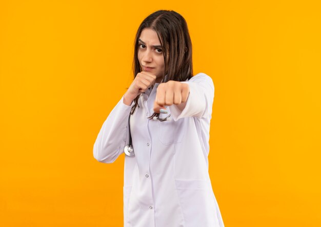 Giovani donne medico in camice bianco con uno stetoscopio intorno al collo guardando in avanti con la faccia seria in posa come un pugile in piedi sopra la parete arancione