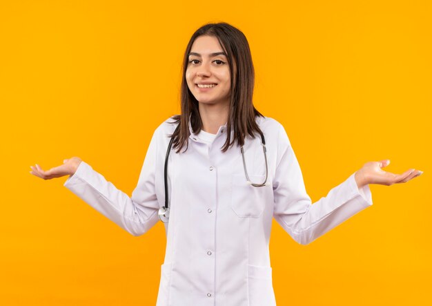 Giovani donne medico in camice bianco con uno stetoscopio intorno al collo guardando in avanti confuso, sorridente diffondendo le braccia ai lati in piedi sopra la parete arancione