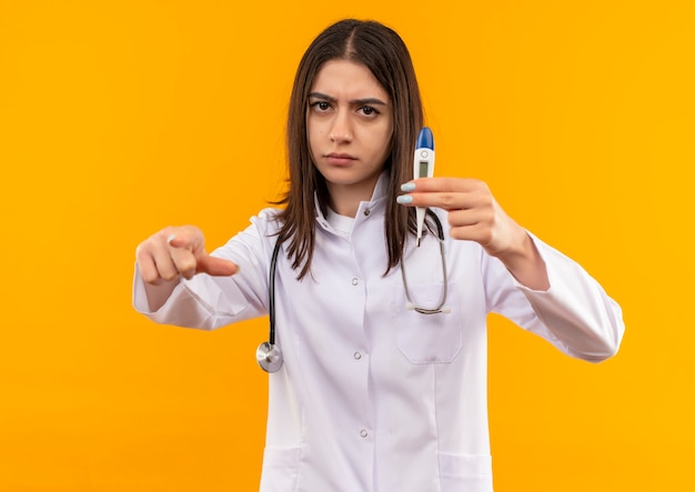 Giovani donne medico in camice bianco con uno stetoscopio intorno al collo tenendo il termometro digitale che punta con il dito indice in avanti con la faccia seria in piedi sopra la parete arancione