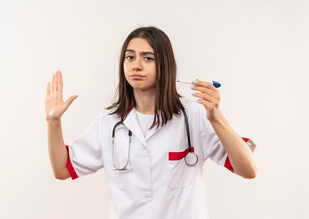 Giovani donne medico in camice bianco con uno stetoscopio intorno al collo tenendo il termometro digitale facendo il segnale di stop con la mano con la faccia seria in piedi sopra il muro bianco