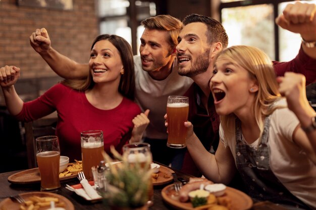 Giovani eccitati che guardano una partita di sport in TV mentre bevono birra e mangiano in un pub