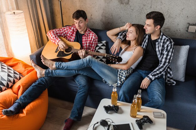 Giovani felici che si divertono, amici festeggiano a casa, compagnia hipster insieme, due uomini una donna, suonare la chitarra, sorridente, positivo, rilassato, bere birra